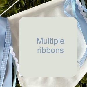 May include: A white canvas banner with the text "Multiple ribbons" printed in blue. The banner has blue and white gingham ribbon, blue satin ribbon, and white scalloped ribbon attached to it.