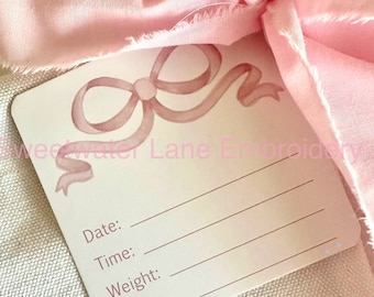 May include: A white card with a pink watercolor bow design. The card has the words "Date:", "Time:", "Weight:", and "Length:" printed on it.