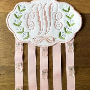 K&ouml;nnte beinhalten: Ein wei&szlig;er Stoffoval mit einem rosafarbenen Monogramm "E.M.E." umgeben von gr&uuml;nen Bl&auml;ttern und blauen Bl&uuml;ten. Der Oval ist an f&uuml;nf rosa B&auml;nder mit durchsichtigen Plastikclips befestigt.