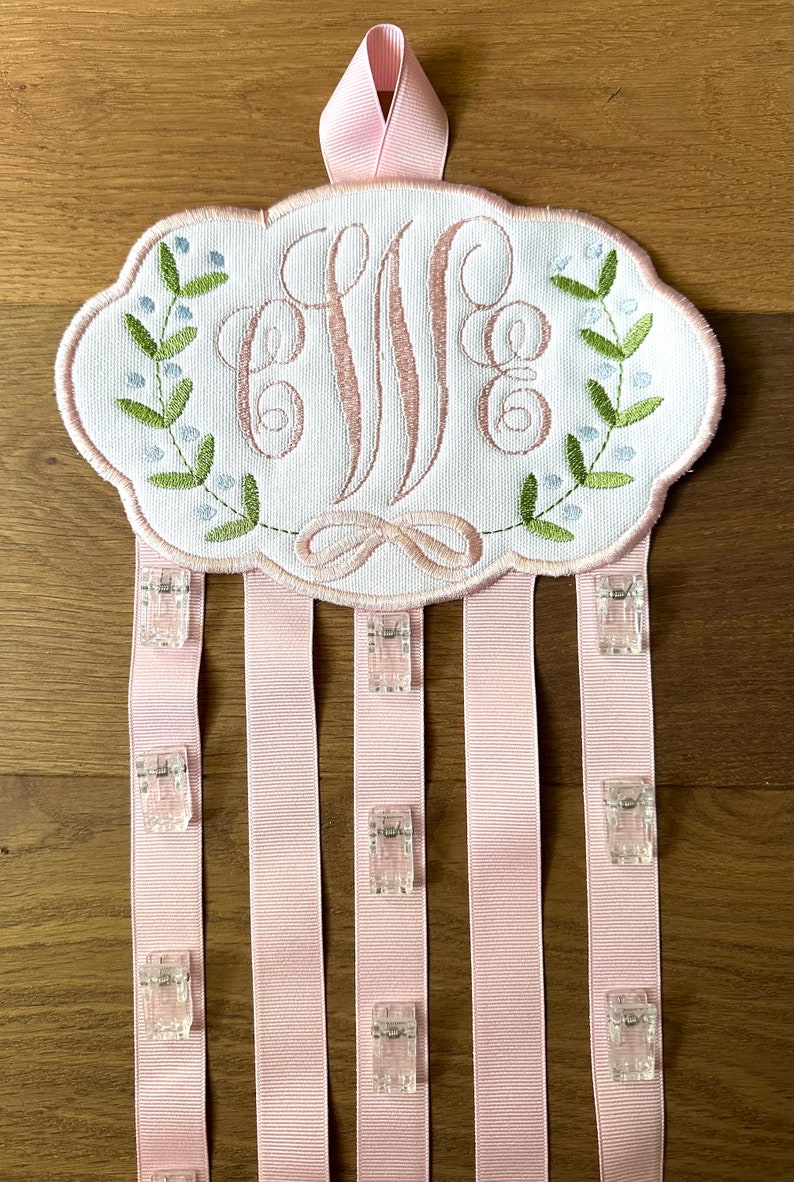 K&ouml;nnte beinhalten: Ein wei&szlig;er Stoffoval mit einem rosafarbenen Monogramm "E.M.E." umgeben von gr&uuml;nen Bl&auml;ttern und blauen Bl&uuml;ten. Der Oval ist an f&uuml;nf rosa B&auml;nder mit durchsichtigen Plastikclips befestigt.