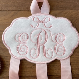 K&ouml;nnte beinhalten: Ein wei&szlig;er Stoff-Haarbogenhalter mit einer rosa Schleifen&ouml;se oben. Der Halter hat ein rosa besticktes Monogramm "ERE" und vier rosa B&auml;nder, die herunterh&auml;ngen.