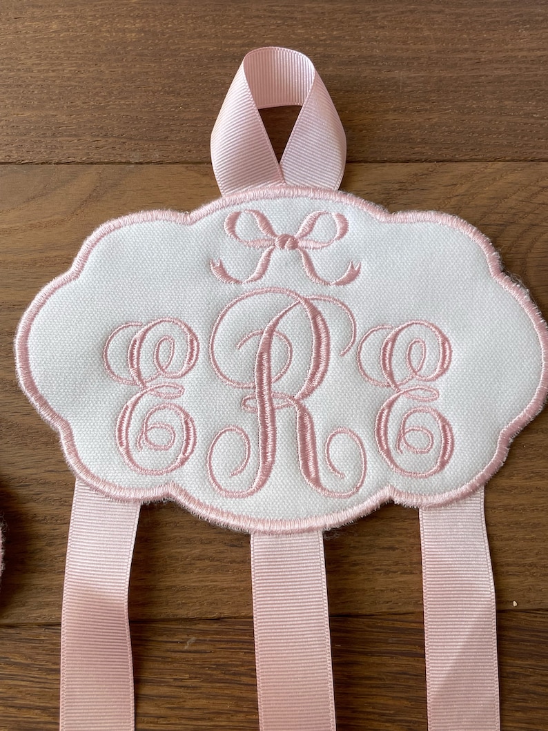 K&ouml;nnte beinhalten: Ein wei&szlig;er Stoff-Haarbogenhalter mit einer rosa Schleifen&ouml;se oben. Der Halter hat ein rosa besticktes Monogramm "ERE" und vier rosa B&auml;nder, die herunterh&auml;ngen.
