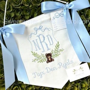 May include: A white banner with blue ribbon ties, embroidered with the initials "NRD" in a blue script font, a leafy wreath, and a brown teddy bear. The banner also includes the name "Nyx Don Ryals" in blue script font.