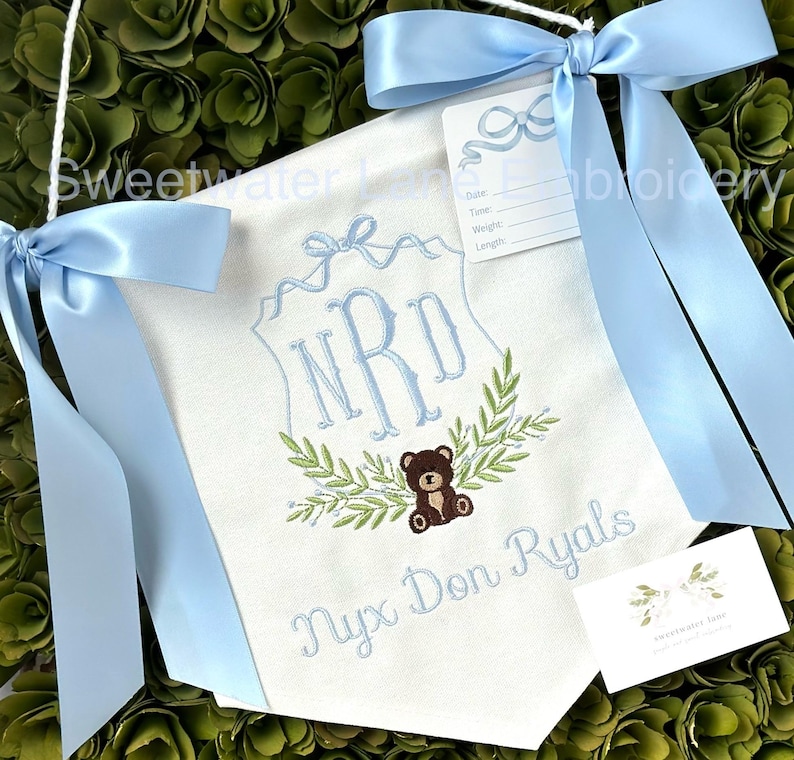 May include: A white banner with blue ribbon ties, embroidered with the initials "NRD" in a blue script font, a leafy wreath, and a brown teddy bear. The banner also includes the name "Nyx Don Ryals" in blue script font.