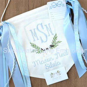 May include: A white banner with blue ribbons and a blue embroidered monogram "MSN" with a wreath of leaves and a dog. The banner also includes the text "Mason Neal Silveia".