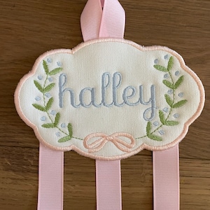 K&ouml;nnte beinhalten: Ein wei&szlig;er Stoff-Haarbandhalter mit dem Namen "halley" in blau gestickt. Der Halter hat eine rosa Schleife zum Aufh&auml;ngen und drei rosa B&auml;nder zum Halten von Schleifen.