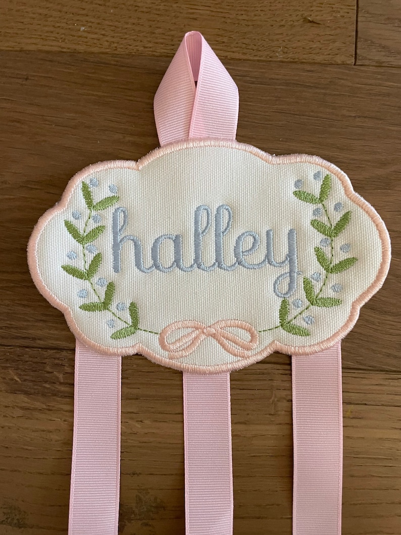 K&ouml;nnte beinhalten: Ein wei&szlig;er Stoff-Haarbandhalter mit dem Namen "halley" in blau gestickt. Der Halter hat eine rosa Schleife zum Aufh&auml;ngen und drei rosa B&auml;nder zum Halten von Schleifen.