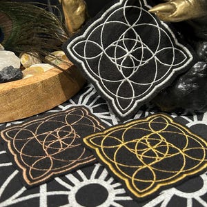 Puede incluir: Tres parches cuadrados negros con un diseño de nudo celta blanco, dorado y bronce. Los parches están bordados con un patrón detallado e entrelazado. Se muestran sobre una superficie estampada en blanco y negro.