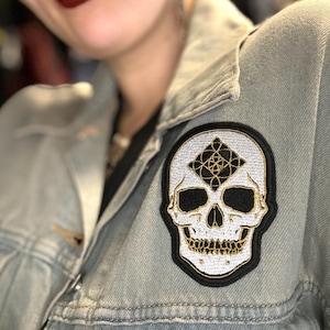 May include: A white skull embroidered patch with a black outline and a gold geometric design in the eye sockets. The patch is sewn onto a light blue denim jacket.