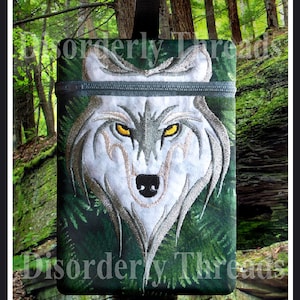 May include: A green zippered pouch with a white wolf embroidered on the front. The wolf has yellow eyes and a black nose. The pouch is surrounded by a green forest background.