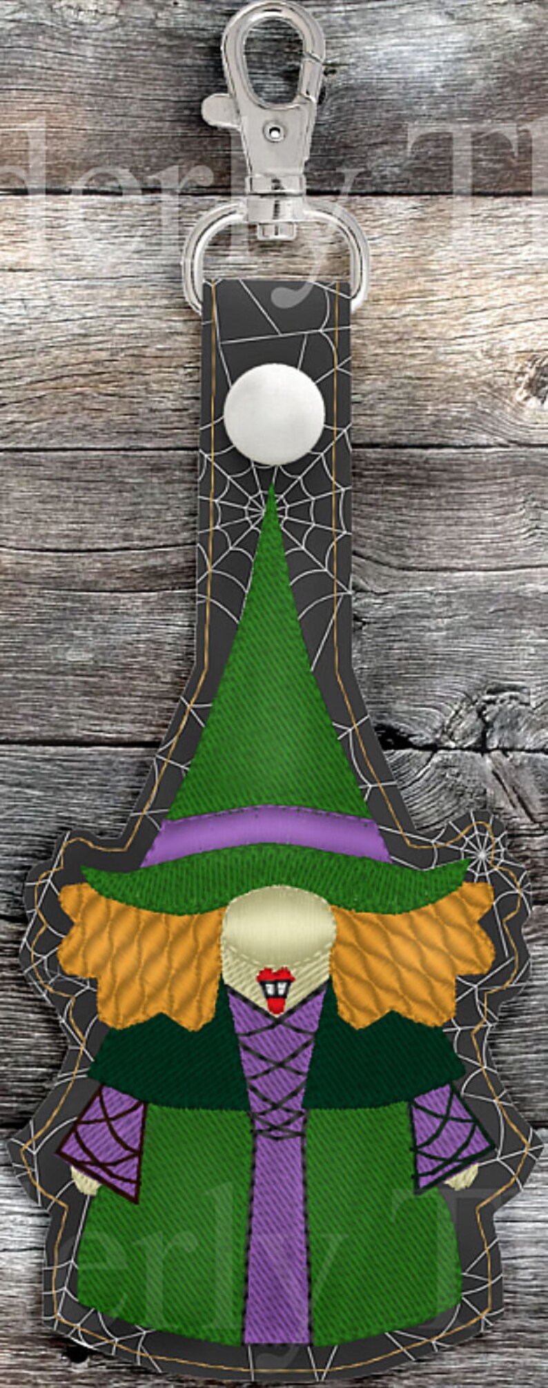 May include: A witch key chain with a green hat, purple dress, and orange hair. The witch is surrounded by a black spider web design.