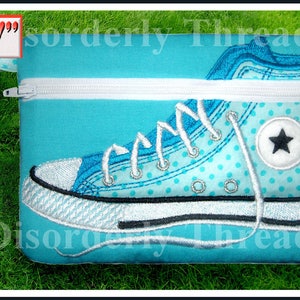 Puede incluir: Una bolsa de cremallera azul turquesa con una cremallera blanca y un diseño bordado blanco y azul de una zapatilla con una estrella blanca en el lateral. La bolsa tiene una correa de lunares azul turquesa. La bolsa mide 12,7x17,8 cm.