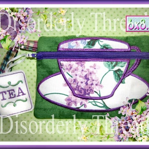 May include: A green zippered pouch with a purple zipper and a teacup design. The teacup is white with a purple floral pattern and a purple outline. The pouch is 6x8.5 inches and has a tag that says "TEA".