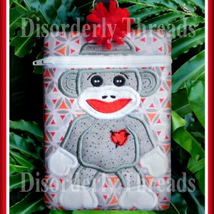 May include: A gray sock monkey with a red heart on its chest is embroidered on a zippered pouch with a red pom-pom on top. The pouch has a geometric pattern of orange, white, and gray.