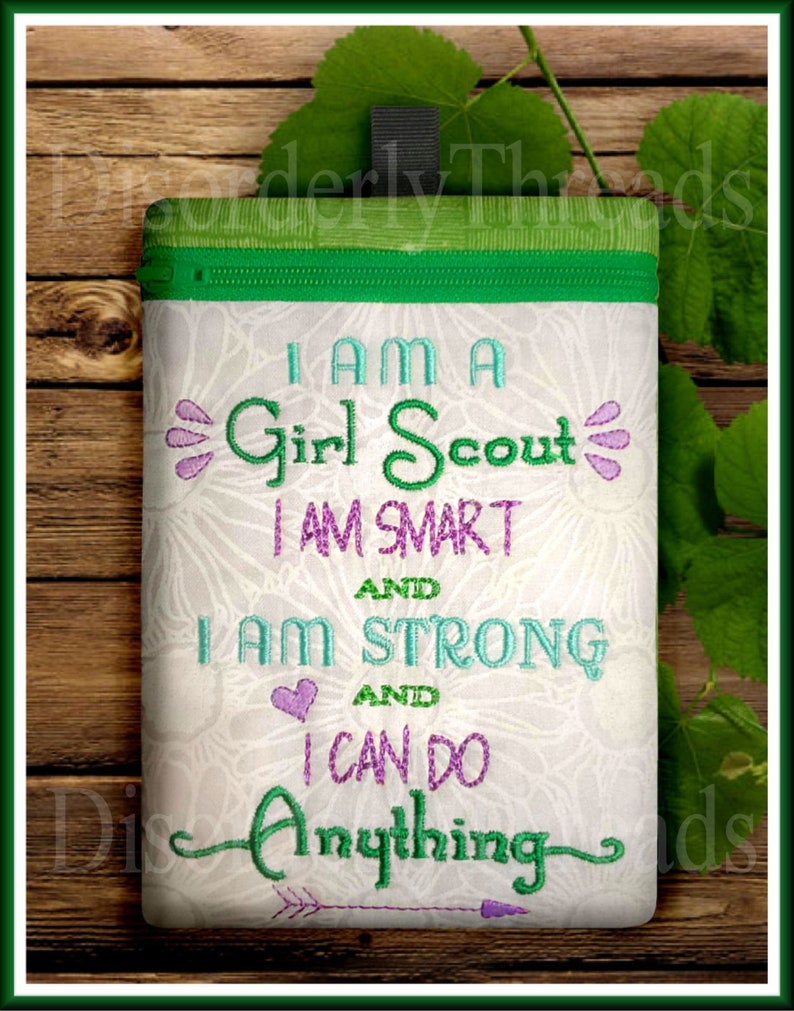 Puede incluir: Una bolsa blanca con cremallera, con la parte superior verde y un dise&ntilde;o bordado en verde y morado que dice "Soy una Girl Scout, soy inteligente y soy fuerte y puedo hacer cualquier cosa".