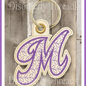 May include: Purple embroidered letter M keychain with a white background and a gold ring. The letter M is in a cursive font and has a pattern of small circles inside.