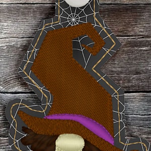 May include: A witch gnome keychain with a brown hat, purple dress, and orange and brown accents. The gnome is on a black background with a white spiderweb design.