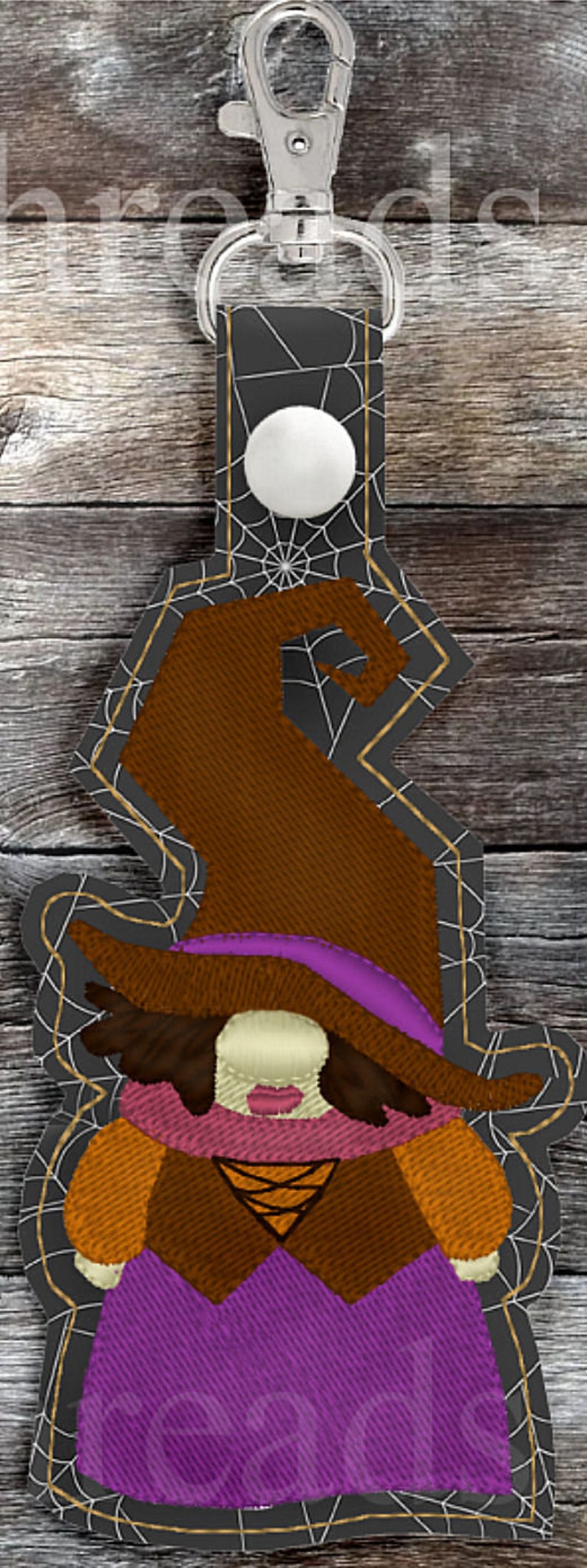 May include: A witch gnome keychain with a brown hat, purple dress, and orange and brown accents. The gnome is on a black background with a white spiderweb design.