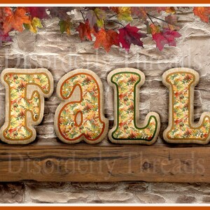 May include: A wooden sign with the word "FALL" spelled out in large letters. The letters are covered in a fabric with a fall leaf pattern in shades of orange, yellow, and green. The sign is sitting on a wooden shelf against a stone wall.