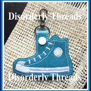 May include: Teal blue fabric shoe keyring with white stitching and a white button. The keyring has a silver metal clasp. The text "Disorderly Threads" is visible on the fabric.