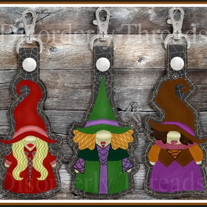 May include: Three Halloween witch keychains with different colored hats and outfits. The witches are wearing red, green, and brown hats with purple dresses and black spiderweb accents.