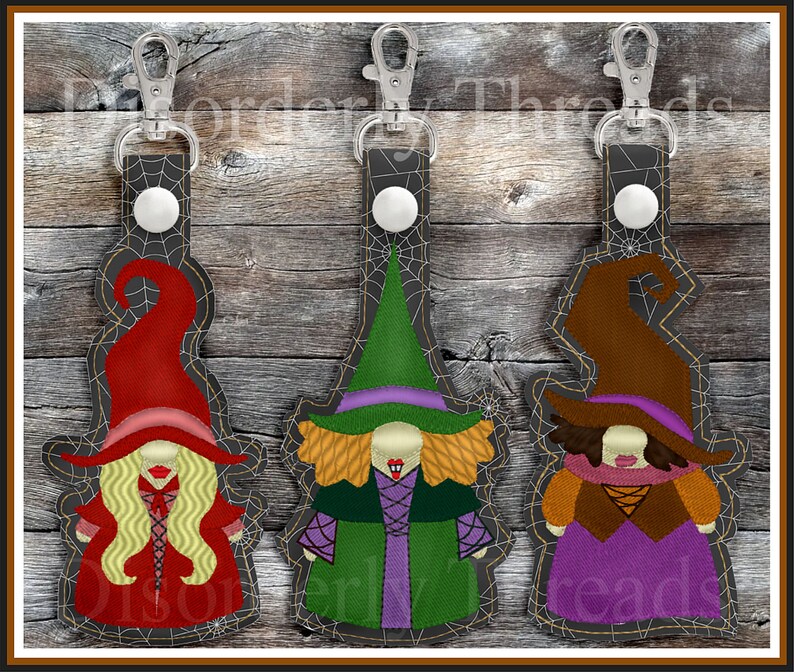 May include: Three Halloween witch keychains with different colored hats and outfits. The witches are wearing red, green, and brown hats with purple dresses and black spiderweb accents.