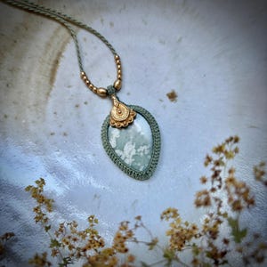Op de afbeelding: Een druppelvormige hanger met een gespikkelde saliegroene steen in een geweven saliegroen koordframe. De hanger is bekroond met een decoratief goudkleurig metalen element en hangt aan een bijpassende koordketting die is versierd met goudkleurige kralen.