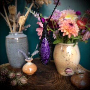 May include: A purple and black gemstone pendant necklace with a black cord. The pendant is a teardrop shape and is wrapped in black cord.