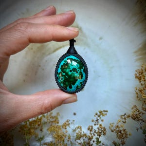 Op de afbeelding: Een ovaalvormige hanger met een gepolijste, donkergroene en blauwgroene steen met cirkelvormige patronen die aan malachiet doet denken. De steen is gevat in een zwarte macramé koordzetting met een lusje aan de bovenkant voor een ketting.