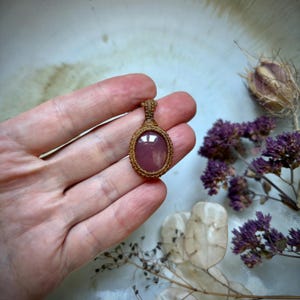 Op de afbeelding: Een handgemaakte hanger met een gepolijste, ovale, mauve edelsteen gezet in een macrame frame. De hanger hangt aan een geweven koord, wat een natuurlijke, aardse uitstraling geeft. De achtergrond bevat gedroogde bloemen en bladeren.