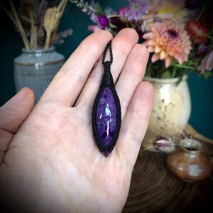 May include: A purple and black gemstone pendant with a black cord. The pendant is in the shape of a teardrop and is wrapped in black cord.