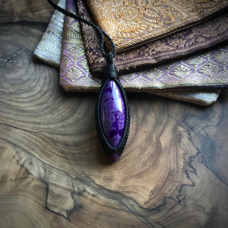 May include: A purple and white agate gemstone pendant with a black cord. The pendant is wrapped in black cord and has a teardrop shape.