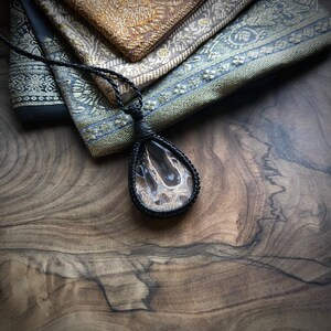 Op de afbeelding: Een zwarte koordketting met een traanvormige hanger met een bruine en witte agaat steen.