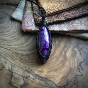 May include: A purple and white gemstone pendant with a black cord. The pendant is wrapped in a black cord and has a unique, marbled pattern.