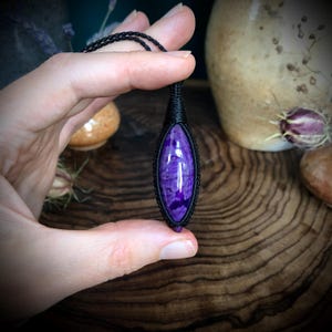 May include: A purple and white gemstone pendant with a black cord. The pendant is wrapped in black cord and has a teardrop shape.