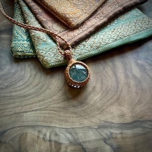 Puede incluir: Un colgante de piedra preciosa verde translúcida y redonda, engastado en un marco de cordón de macramé color canela. El colgante cuelga de un cordón trenzado a juego, con un intrincado trabajo de nudos que asegura la piedra en su marco.