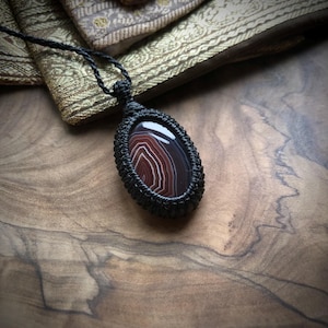 Op de afbeelding: Een handgemaakte macramé ketting met een ovale agaat hanger. De steen toont een patroon van rode, witte en bruine banden. De hanger is omhuld met een zwarte macramé weefsel en hangt aan een zwart koord.