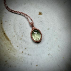 Pode incluir: Um colar com pendente apresentando uma pedra preciosa oval, verde-clara translúcida, com inclusões escuras em forma de agulha. A pedra está envolvida numa armação de cordão tecido rosa-pálido que continua para cima formando um cordão fino.