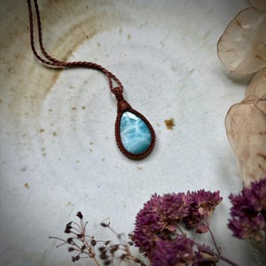 Op de afbeelding: Een handgemaakte macramé-ketting met een druppelvormige larimarsteen. De steen vertoont een lichtblauw en wit patroon, ingekapseld in een bruin macramé-weefsel. De ketting heeft een bruin koord.