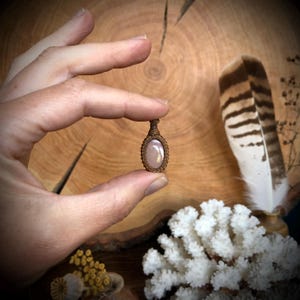 Op de afbeelding: Een kleine, handgemaakte hanger met een lichtroze edelsteen, gezet in een bruine macramé-omgeving. De hanger wordt vastgehouden, waarbij het ingewikkelde ontwerp wordt benadrukt. De achtergrond bevat een houten oppervlak, een veer en wit koraal.