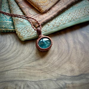 May include: A pendant necklace featuring a round, translucent teal-colored stone encased in a brown macrame cord setting. The cord is woven into a decorative knot pattern around the stone, with a matching brown cord extending upward.