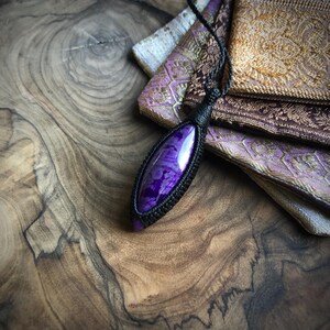 May include: A purple and black gemstone pendant with a black cord. The pendant is on a wooden surface with a patterned fabric in the background.