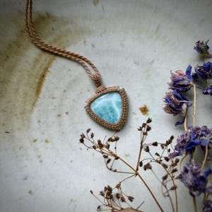 May include: A triangular pendant necklace featuring a light blue gemstone, set in a macrame design. The necklace has a tan cord and is displayed on a neutral surface with dried flowers.