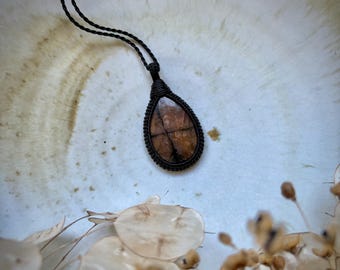 May include: A teardrop-shaped pendant necklace featuring a brown stone with a cross-like pattern, encased in a black woven macrame setting. The necklace has a black cord. The background is a light-colored, textured surface.