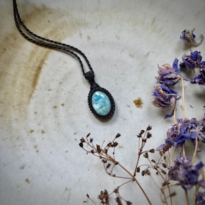Op de afbeelding: Handgemaakte macramé ketting met een lichtblauwe ovale edelsteen hanger. De hanger is gevat in een zwarte macramé weefsel, hangend aan een bijpassend zwart koord. Gedroogde paarse bloemen en bruine stelen zijn rond de ketting gerangschikt.