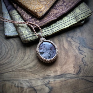 Op de afbeelding: Een close-up van een ketting met een hanger van donkerpaarse en grijze steen, gezet in een geweven macramé frame. De ketting ligt op een houten oppervlak.