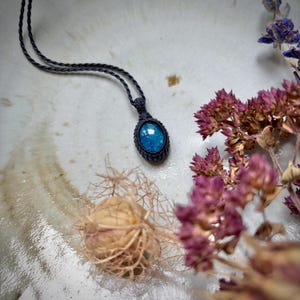 May include: A pendant necklace featuring a polished, oval-shaped blue gemstone encased in a dark, intricately knotted macramé setting. The pendant hangs from a thin, matching dark cord, showcasing a vibrant, reflective blue surface texture.