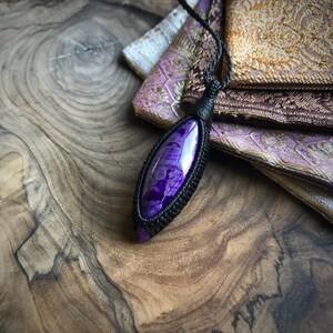 May include: A purple and black gemstone pendant necklace with a knotted cord. The pendant is a long oval shape and has a shiny, polished finish.