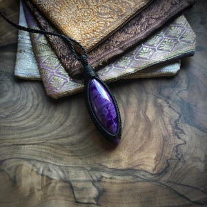 May include: A purple and white agate gemstone pendant on a black cord necklace. The pendant is wrapped in black cord and has a teardrop shape.