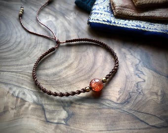 May include: A brown macrame bracelet with a central orange-red bead and small gold-colored accents. The bracelet is adjustable and sits on a wooden surface. The style is bohemian and handcrafted.